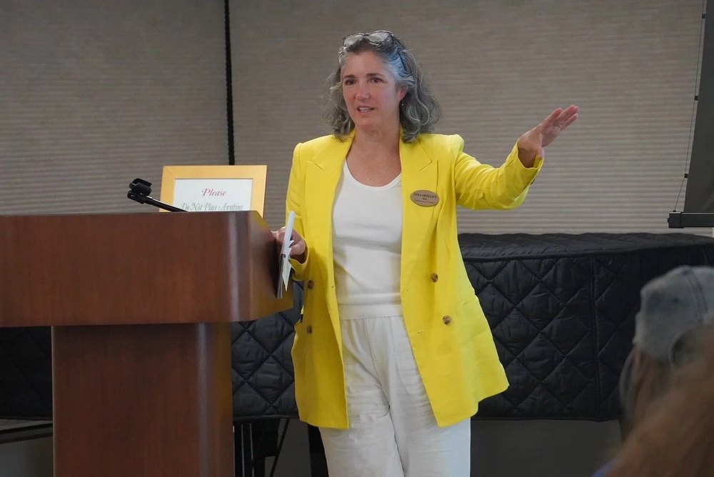 Gina DeFelice speaking at a podium in a community room, wearing a yellow blazer and a campaign name tag